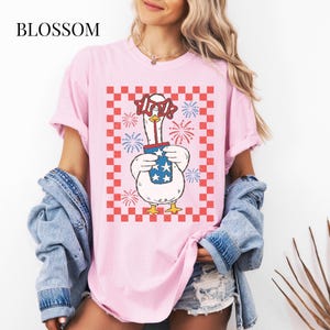 May include: Pink t-shirt with a graphic of a goose wearing star-shaped glasses and holding a firework container with stars. The design is set against a red and white checkered background, with the word "BLOSSOM" at the top left.