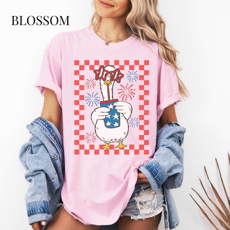May include: Pink t-shirt with a graphic of a goose wearing star-shaped glasses and holding a firework container with stars. The design is set against a red and white checkered background, with the word "BLOSSOM" at the top left.