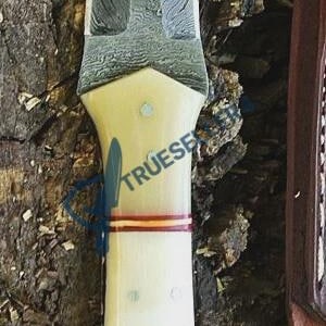 May include: A Damascus steel dagger with a cream-colored handle, red and gold accents, and decorative rivets. The blade has a unique pattern. The dagger is displayed on a textured wooden surface.