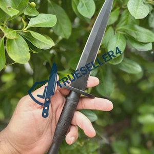 Puede incluir: Una daga negra con una hoja puntiaguda y un mango texturizado. La daga tiene una guarda y un pomo. El texto "TRUESELLER4" es visible en la hoja. La daga se muestra sobre un fondo de follaje verde.