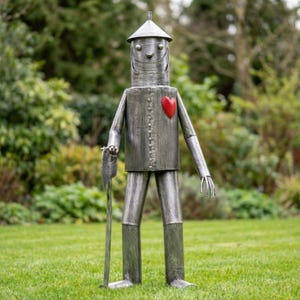 May include: A metal sculpture of the Tin Man from The Wizard of Oz, standing on grass. The sculpture is silver with a red heart and holds an axe. The Tin Man wears a pointed hat and has a smiling face.