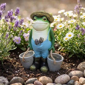 May include: A whimsical garden frog statue wearing blue overalls, a straw hat, and black boots, holding two gray buckets with rope handles. The frog has a friendly expression and is surrounded by flowers.