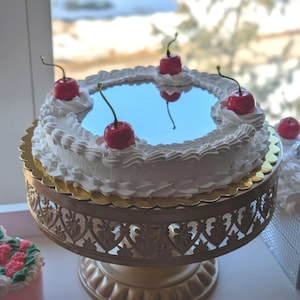 May include: A white frosted cake with red cherries on top, presented on a gold-colored decorative cake stand. The cake has a mirrored center and is surrounded by whipped cream. The stand has a detailed, openwork design.