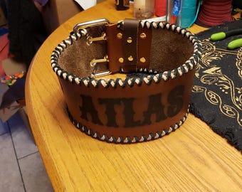 Puede incluir: Un cinturón de levantamiento de pesas de cuero marrón con costuras blancas y un logotipo "ATLAS" negro. El cinturón tiene una hebilla plateada y está sobre una superficie de madera.