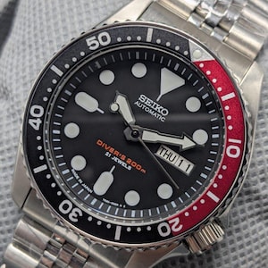 Puede incluir: Reloj de buceo automático Seiko de acero inoxidable con esfera negra, marcadores luminosos blancos y visualización de la fecha. El bisel giratorio tiene secciones negras y rojas. La pulsera es de acero inoxidable.
