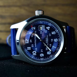 May include: A silver wristwatch with a dark blue face and a blue leather strap. The watch face displays white numerals and hands, with the brand name "HAMILTON" visible. The watch has a date display at the 3 o'clock position.