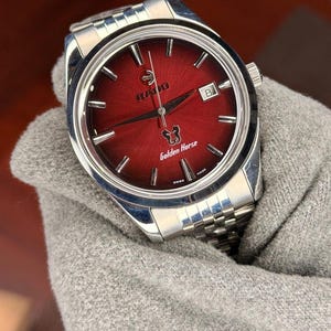 May include: A stainless steel Rado Golden Horse watch with a red dial. The watch features a date window at the 3 o'clock position and a silver-tone bracelet. The dial has the Rado logo and the words "Golden Horse" printed on it.