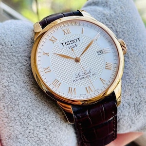 以下が含まれることがあります： 白い文字盤とローマ数字のインデックスを備えた、ゴールドトーンのTissot Le Locle Powermatic 80腕時計。3時位置に日付表示があり、茶色の革ストラップが付いています。グレーの表面に表示されています。