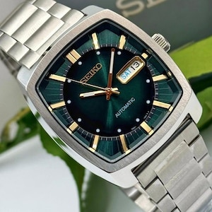 May include: A silver-toned Seiko automatic wristwatch with a square green face. The watch features gold-colored hands and markers, a day-date display, and a silver band. The watch face has the word "SEIKO" and "AUTOMATIC" printed on it.