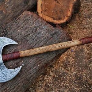 May include: A double-bladed axe with a wooden handle and a silver-colored head. The axe head features an intricate Celtic knot design. The handle is wrapped with brown leather near the head. The axe rests on a weathered wooden surface.