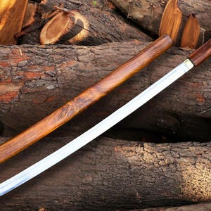 Puede incluir: Una espada katana japonesa con vaina y empuñadura de madera. La hoja es plateada, y la empuñadura y la vaina son de color marrón oscuro. La espada descansa sobre una pila de troncos.