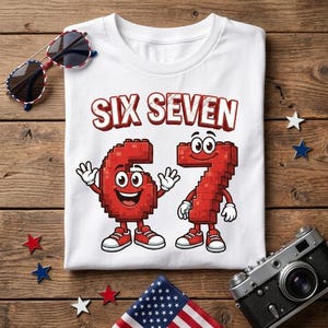 May include: A white t-shirt featuring the text SIX SEVEN in red block letters above two cartoon characters shaped like the numbers six and seven, constructed from red building blocks with white gloves and red sneakers on a wooden surface.