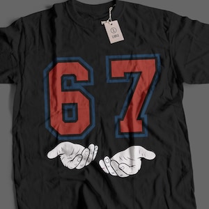 Può includere: T-shirt nero con il numero "67" in rosso con contorno blu e due mani bianche a coppa. Una piccola etichetta è attaccata alla maglietta. La maglietta è taglia Large.
