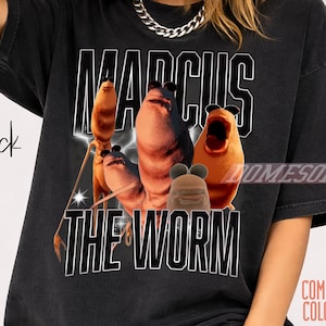 May include: Black t-shirt featuring the text "MARCUS THE WORM" in a white, collegiate-style font. The graphic includes cartoon worms in shades of brown and orange. The shirt is a Comfort Colors brand.