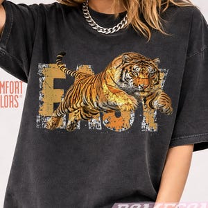 Può includere: T-shirt Comfort Colors grigio scuro con grafica arancione e gialla invecchiata di una tigre che salta sopra la parola "EASY". La tigre ha strisce arancioni e nere. La maglietta ha un aspetto vintage e usurato.