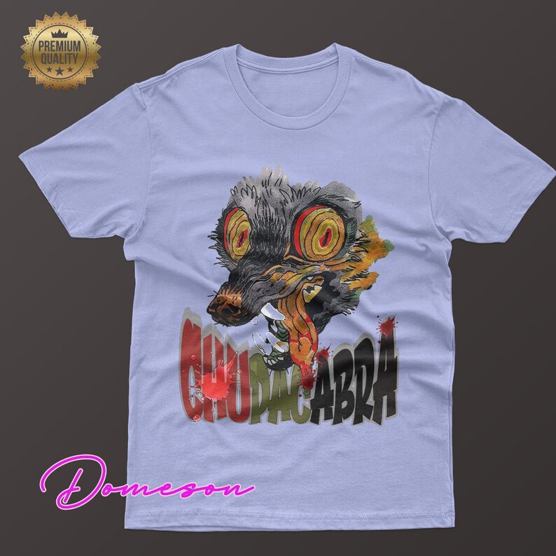 May include: Light blue t-shirt featuring a watercolor illustration of a creature with large, yellow eyes and the word "CHUPACABRA" in red and black. The shirt has a crew neck and short sleeves.