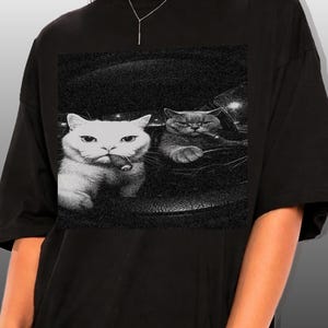 May include: Black t-shirt featuring a black and white graphic of two cats. One cat is white and holding a cigar, while the other is gray. The cats are in a car. The shirt is a crew neck.