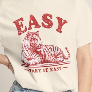 Può includere: T-shirt color crema con grafica rossa di una tigre e la scritta "EASY" sopra. La frase "TAKE IT EASY" è stampata sotto. Un capo casual, ideale per un look rilassato.