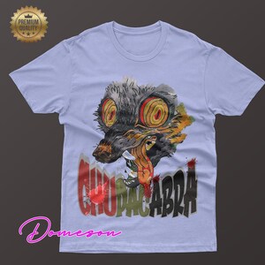 May include: Light blue t-shirt featuring a watercolor illustration of a creature with large yellow eyes and the word "CHUPACABRA" in red and black. The shirt has a crew neck and short sleeves. A "Premium Quality" seal is in the upper left corner.