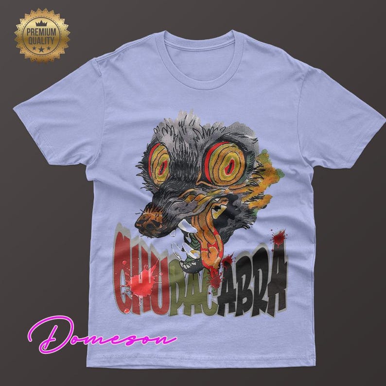 May include: Light blue t-shirt featuring a watercolor illustration of a creature with large yellow eyes and the word "CHUPACABRA" in red and black. The shirt has a crew neck and short sleeves. A "Premium Quality" seal is in the upper left corner.
