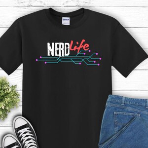 Può includere: Maglietta nera con il testo "NERDlife" in rosso e blu con un design a circuito stampato in blu e rosa.