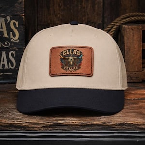 Può includere: Un cappellino da baseball beige con visiera nera e una toppa rettangolare in pelle marrone centrata sul davanti. La toppa presenta un grafico di un teschio di longhorn con il testo ELLA'S FELLAS stampato in modo circolare attorno al teschio.
