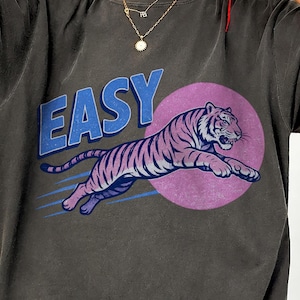 Può includere: T-shirt grigio scuro con una grafica di tigre che salta in rosa e viola, su un cerchio viola. La parola "EASY" è stampata sopra la tigre in blu, in stile vintage.