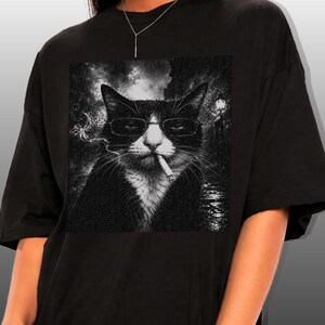 May include: Black t-shirt featuring a black and white graphic of a cat wearing glasses and smoking a cigarette. The cat has black and white fur and is set against a blurred background.