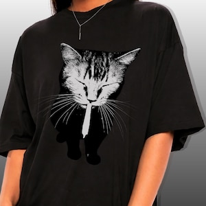 May include: Black t-shirt featuring a grayscale graphic of a cat with closed eyes, holding a white object in its mouth. The cat's fur is detailed with stripes. The shirt has short sleeves and a relaxed fit.