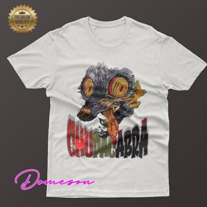May include: White t-shirt featuring a colorful graphic of a creature with large, yellow eyes and the word "CHUPACABRA" in a stylized font. The design includes red, green, and black accents. A "Premium Quality" seal is in the upper left corner.