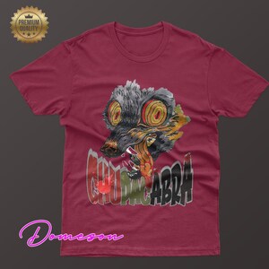 May include: Burgundy t-shirt featuring a graphic of a snarling, multi-colored creature with the word "CHUPACABRA" in a distressed font. The design includes shades of gray, yellow, red, and black. A "Premium Quality" seal is in the upper left.