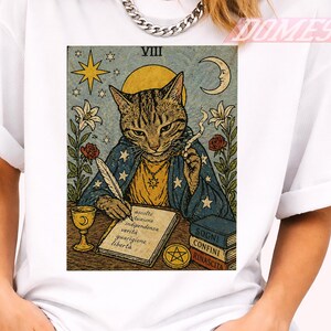 Può includere: T-shirt bianco con un disegno di carta dei tarocchi. La carta raffigura un gatto in una veste blu, che scrive con una penna, con una luna, stelle e fiori sullo sfondo. Il testo sulla carta è in italiano.