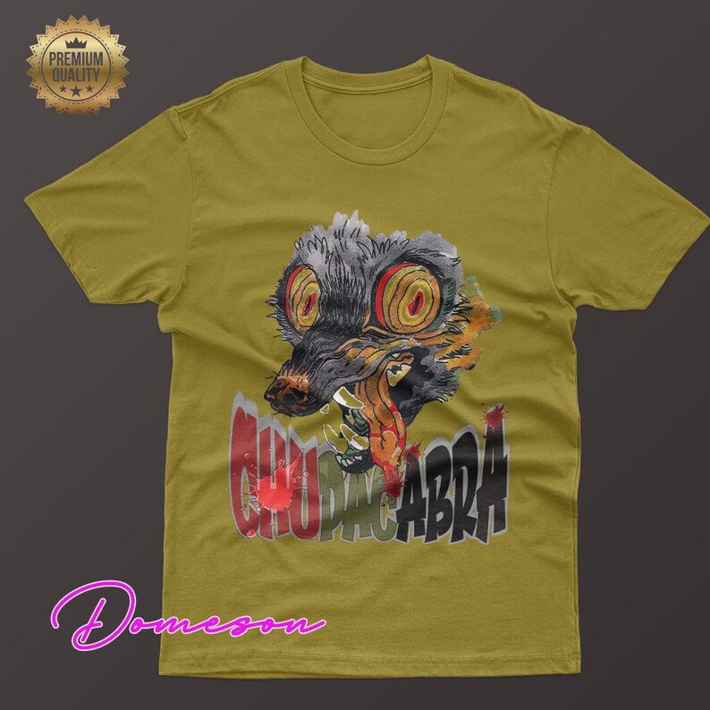 May include: Mustard yellow t-shirt featuring a graphic of a creature with large, red-ringed eyes and the word "CHUPACABRA" in red and black. The shirt has a crew neck and short sleeves. A "Premium Quality" seal is in the upper left corner.