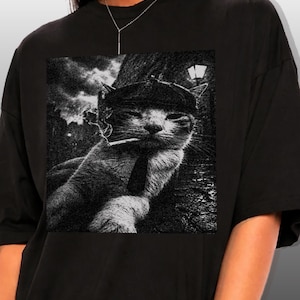 May include: Black t-shirt featuring a black and white graphic of a cat wearing a hat and tie, smoking a cigarette. The cat is set against a backdrop of a street scene with a lamp post and cloudy sky.