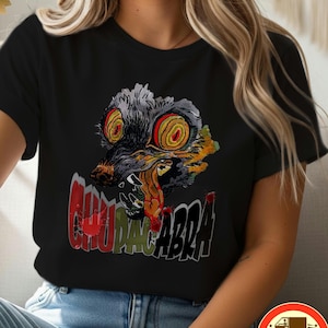 May include: Black t-shirt featuring a colorful graphic of a snarling, wolf-like creature with large, spiral eyes and the word "CHUPACABRA" in red and white text. The design is centered on the front of the shirt.