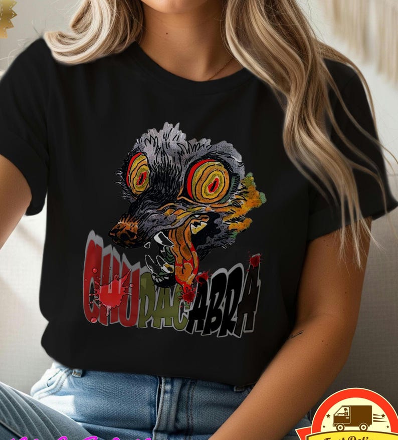 May include: Black t-shirt featuring a colorful graphic of a snarling, wolf-like creature with large, spiral eyes and the word "CHUPACABRA" in red and white text. The design is centered on the front of the shirt.