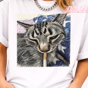 Può includere: T-shirt bianco con un'illustrazione dettagliata di un gatto tabby grigio con una sigaretta in bocca. Il gatto è ambientato su uno sfondo blu e bianco a forma di stella, con un tocco di rosa.