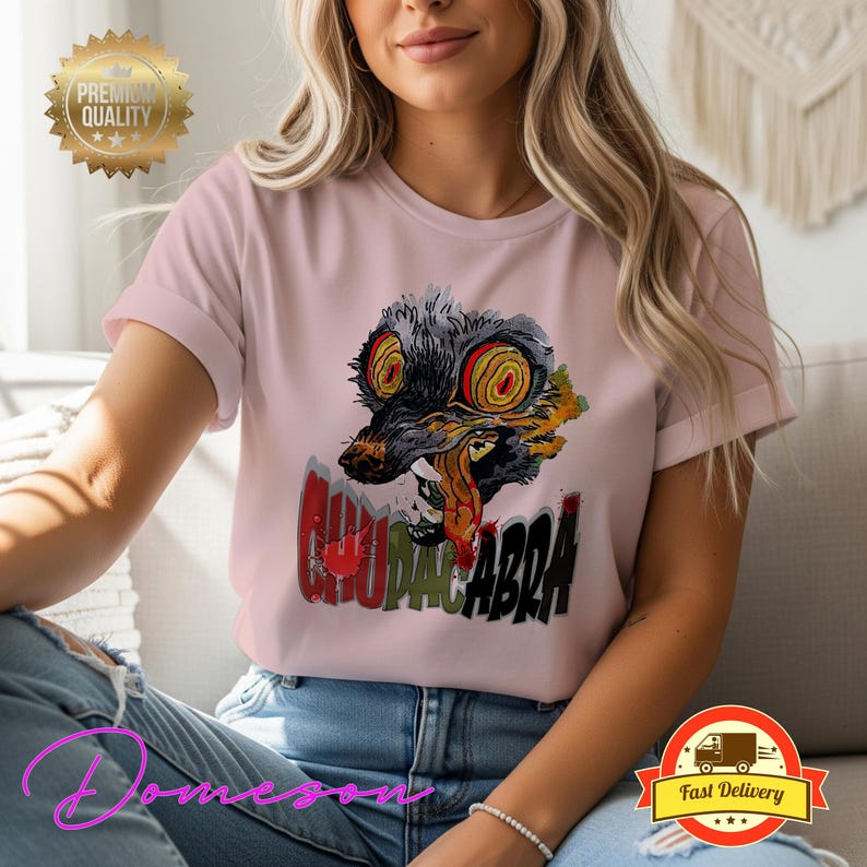 May include: A light pink t-shirt featuring a graphic of a Chupacabra with red, yellow, and black accents. The text "CHUPACABRA" is printed below the graphic in red, green, and black. The shirt is a short-sleeved crew neck.
