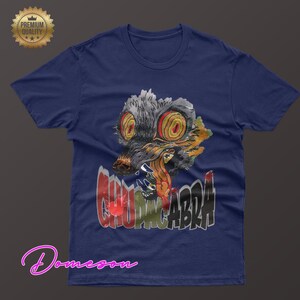 May include: Navy blue t-shirt featuring a graphic of a creature with large yellow eyes and the word "CHUPACABRA" in a graffiti-style font. The design includes red, gray, and orange colors. A "Premium Quality" seal is in the upper left corner.