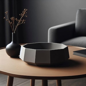 May include: A grey geometric bowl sits on a wooden table with a black vase and dried flowers. The bowl is made of a smooth, matt material and has a unique, modern design.