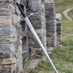 Può includere: Una lunga spada d'argento con un'impugnatura e un design della lama decorativi, appoggiata a un muro di pietra. La spada ha un'impugnatura marrone scuro e una guardia incrociata argentata. La scena è all'aperto con una passerella di legno.