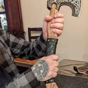 May include: A hand holding a Viking axe with a wooden handle and a leather wrap. The axe head is decorated with intricate carvings and a knotwork design. The axe is approximately 20 inches long.