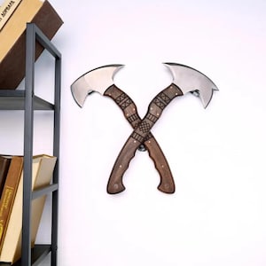 May include: Two crossed axes with polished steel blades and dark wood handles. The handles feature decorative geometric patterns and are mounted on a white wall. The axes are a decorative item.
