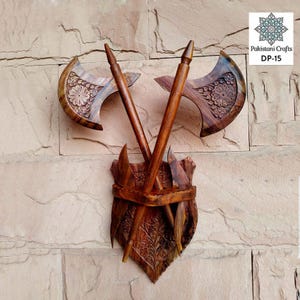 Puede incluir: Una decoración de pared de madera con dos hachas cruzadas con intrincados tallados. Las hachas están montadas en una base con forma de escudo con tallados adicionales. La pieza está etiquetada como "Pakistani Crafts DP-15".