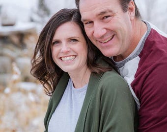 May include: A couple smiling and hugging in a snowy outdoor setting. The woman is wearing a green cardigan over a light blue shirt. The man is wearing a white and red striped shirt.