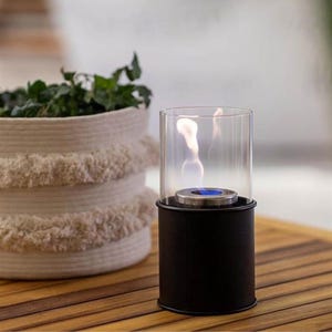 May include: A small, black cylindrical tabletop fireplace with a glass enclosure. The flame is visible inside the glass. A woven basket with greenery is in the background. The fireplace has a silver-colored base and a blue accent.