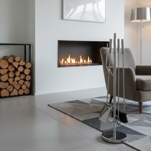 May include: A contemporary living room featuring a fireplace with a flame effect. A set of silver fireplace tools stands in front of a grey armchair. A geometric rug and a stack of logs are also visible. The room has a minimalist design.