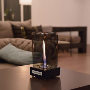 May include: A modern, rectangular tabletop fireplace with a flickering flame. The fireplace is encased in a clear, reflective glass structure with a black base. The name "Michael Gomez" is printed on a small label on the base.