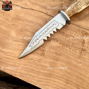 May include: A handcrafted knife with a serrated blade and a light-colored antler handle. The silver blade has etched text and a decorative edge. The knife is displayed on a wooden surface.