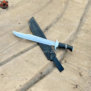 May include: A large, silver blade knife with a black and white handle and a black leather sheath. The knife has a wide, flat blade and a curved guard. The sheath is embossed with a pattern.